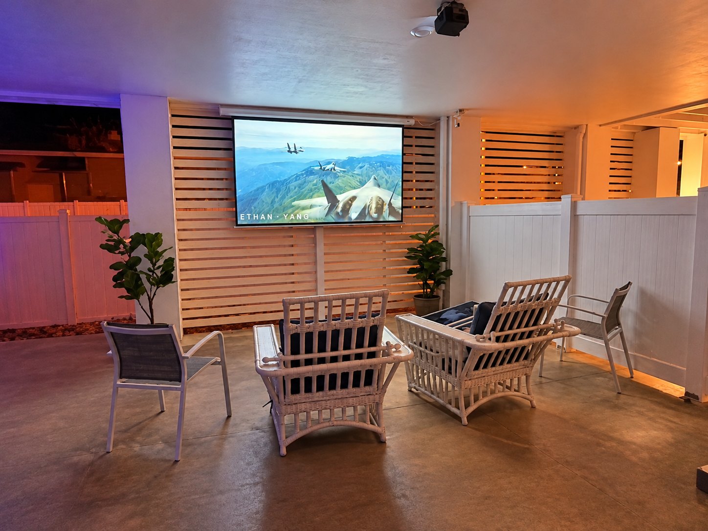 Covered outdoor theater with a large projector screen and white lounge chairs